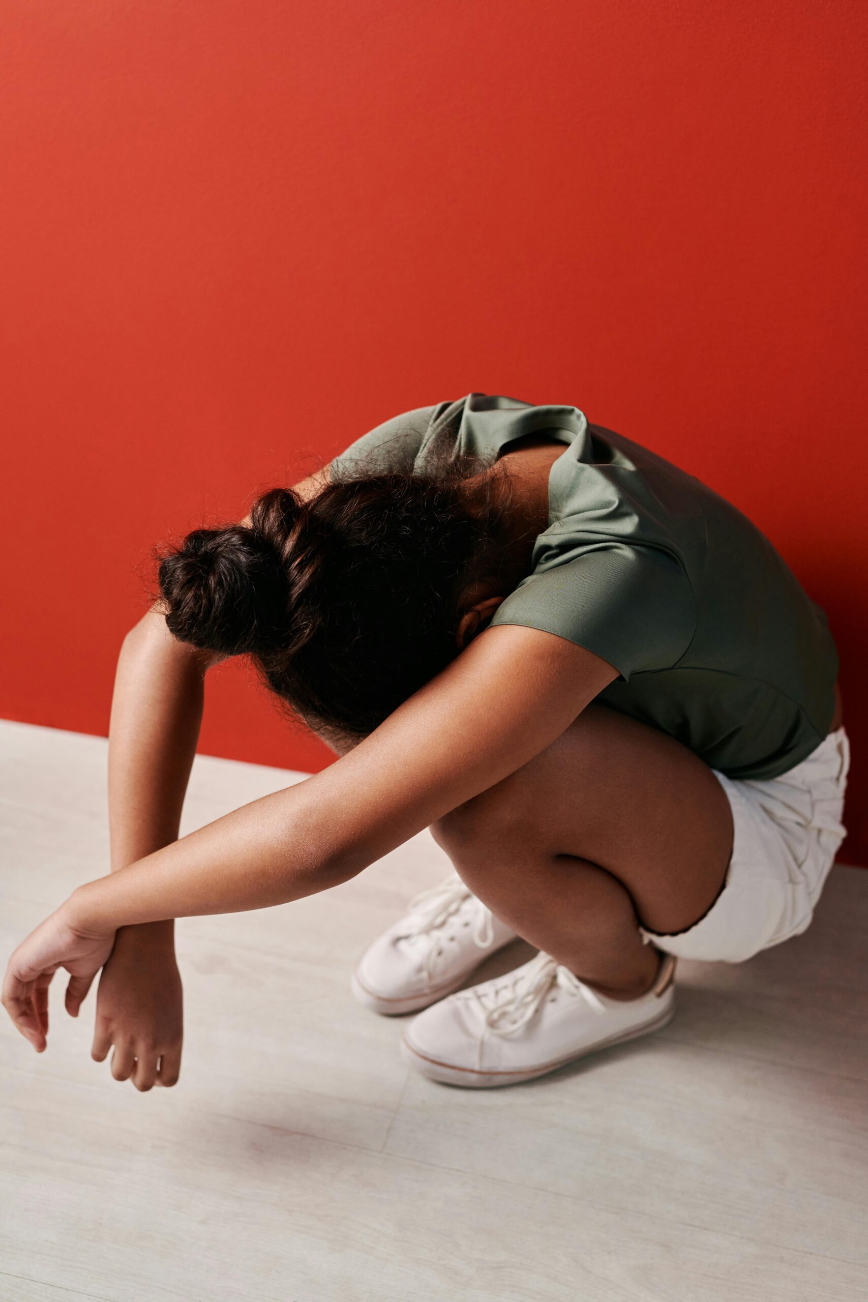 A young girl crouches with her head down wearing white shoes and shorts against a red wall.